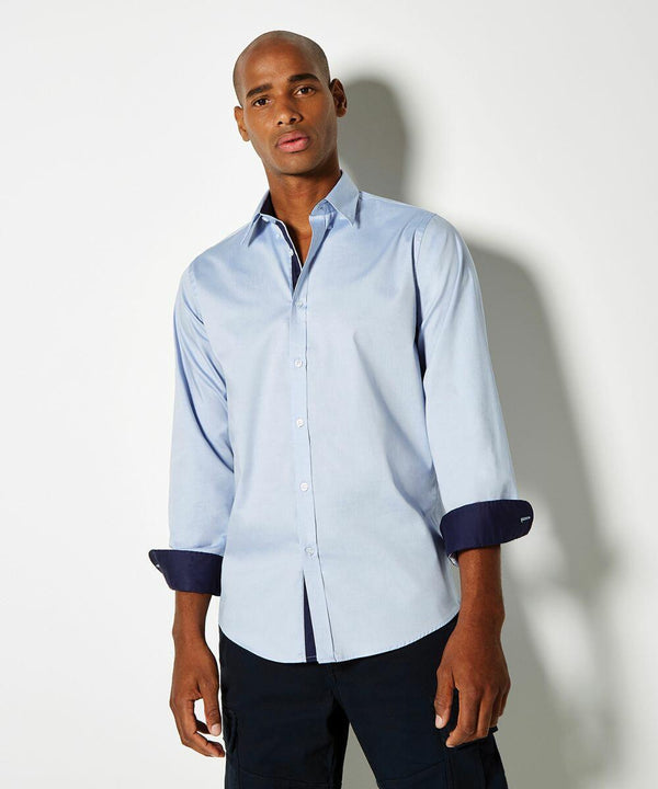 A person stands against a white background wearing a Kustom Kit light blue long-sleeved button-up shirt with dark cuffs and dark pants, gazing directly at the camera.