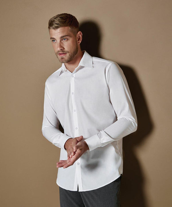 A man with short hair and a beard is wearing a white dress shirt (KK192 by Kustom Kit) and gray pants, standing against a plain brown background. He is looking to the side with his hands brought together.
