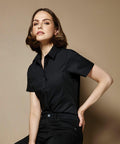 A person with shoulder-length hair wearing a short-sleeve black KK241 shirt from Kustom Kit and black pants, posing against a beige background.
