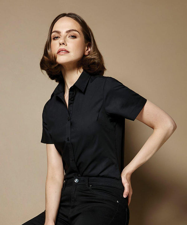 A person with shoulder-length hair wearing a short-sleeve black KK241 shirt from Kustom Kit and black pants, posing against a beige background.