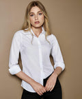 A person with blonde hair wearing a white button-up Kustom Kit KK242 shirt and black pants stands against a brown background.