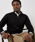 A man sits on a wooden chair wearing a Kustom Kit Regular fit Arundel 1/4-zip jumper over a white collared shirt and khaki trousers, looking directly at the camera with a neutral expression.