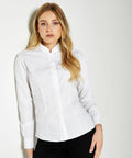 A woman with long blonde hair and a neutral expression is wearing a white collared shirt (KK261 by Kustom Kit) and black pants, standing against a plain light-colored background.