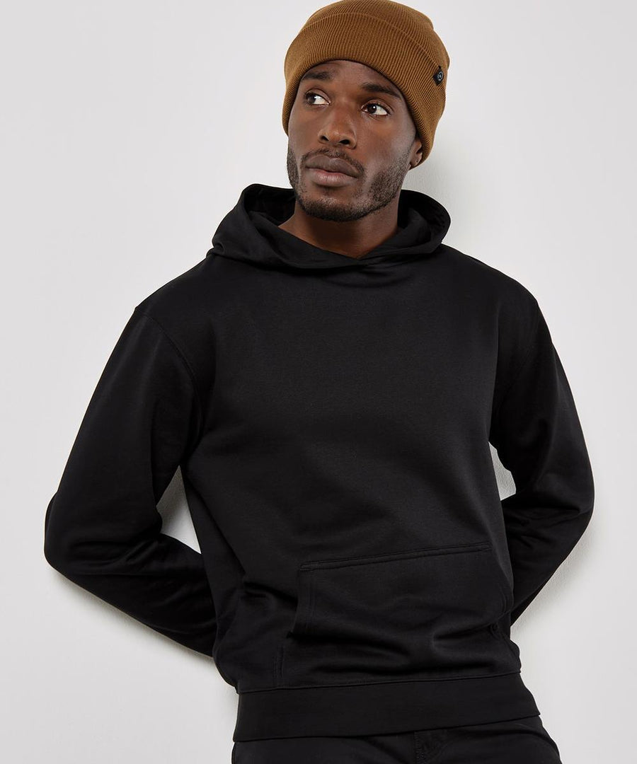 A man wearing a Kustom Kit Regular fit Superwash 60 hoodie in black and a brown beanie stands against a white background, gazing slightly upwards to the left with his hands behind his back.