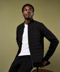 A man in a white T-shirt and the Kustom Kit Regular Fit Quilted Full-Zip Sweatshirt sits on a stool against an olive green background, gazing upwards and to the right.