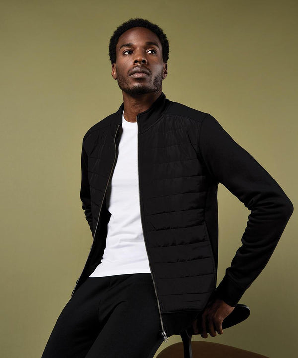 A man in a white T-shirt and the Kustom Kit Regular Fit Quilted Full-Zip Sweatshirt sits on a stool against an olive green background, gazing upwards and to the right.