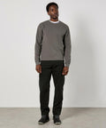 A man stands indoors against a plain light wall, wearing the Kustom Kit Regular Fit Piqué Sweatshirt. He faces slightly left with relaxed arms by his sides.
