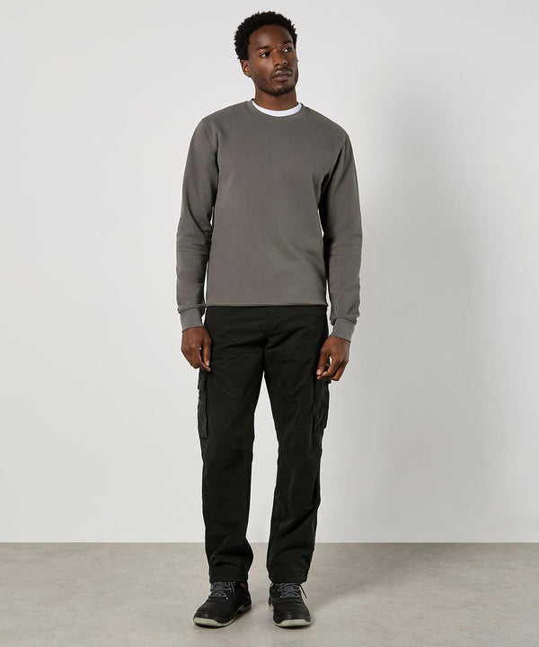 A man stands indoors against a plain light wall, wearing the Kustom Kit Regular Fit Piqué Sweatshirt. He faces slightly left with relaxed arms by his sides.
