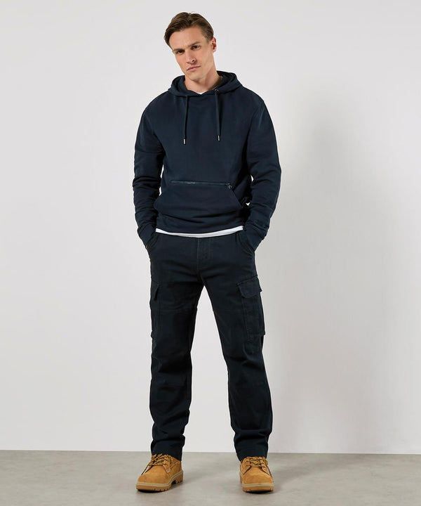 A man stands against a plain white background wearing the Kustom Kit Regular Fit Piqué Hoodie with Zipped Pocket, paired with dark cargo trousers and tan work boots. His hands are in his pockets, and he looks straight ahead with a neutral expression.