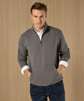 A man wears a Kustom Kit Regular fit 1/4-zip piqué sweatshirt in grey over a white collared shirt, paired with dark blue jeans, standing against a light wooden background with his hands in his pockets.