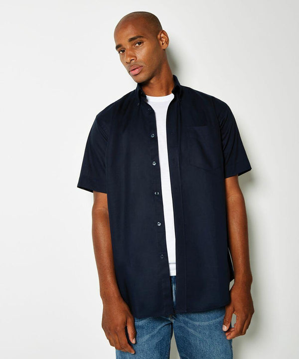 A person wearing a navy blue Kustom Kit KK350 short-sleeve button-down shirt over a white t-shirt stands against a plain white background.
