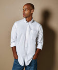 A man stands against a beige background wearing a white KK351 button-up shirt by Kustom Kit and blue jeans, with his hands in his pockets, looking to the side.