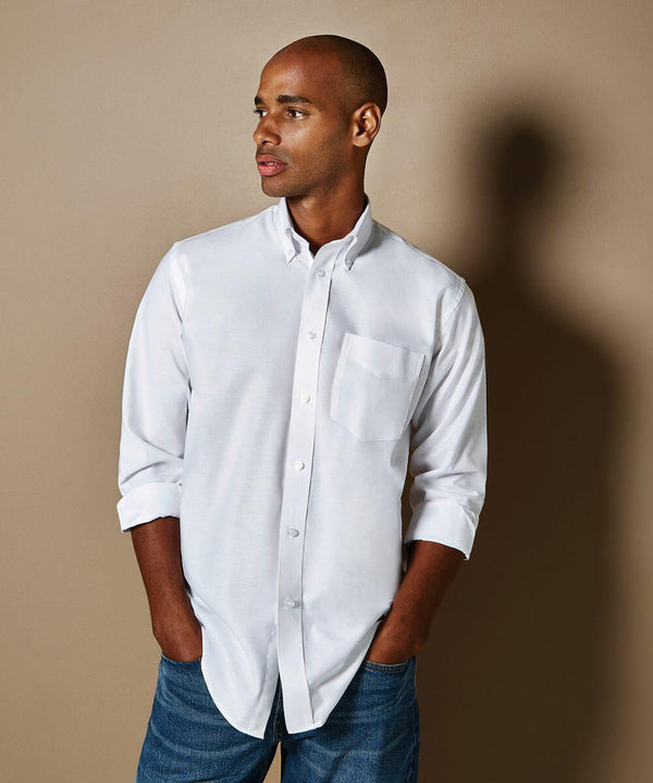 A man stands against a beige background wearing a white KK351 button-up shirt by Kustom Kit and blue jeans, with his hands in his pockets, looking to the side.