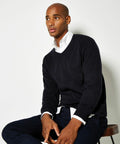 A person with a neutral expression sits on a stool, wearing a Kustom Kit KK352 black V-neck sweater over a white collared shirt and dark pants.