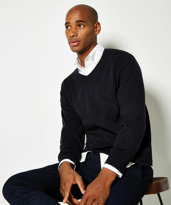 A person with a neutral expression sits on a stool, wearing a Kustom Kit KK352 black V-neck sweater over a white collared shirt and dark pants.