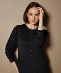 A person with shoulder-length hair, wearing a black button-up Kustom Kit KK355 sweater, stands against a beige background and touches their head with one hand.