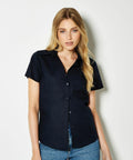 A person with long blonde hair is wearing a short-sleeved, button-up, black KK360 shirt by Kustom Kit and blue jeans, standing against a plain white background.