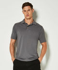 A man wearing a gray KK400 polo shirt by Kustom Kit and dark pants stands against a plain white background, with one hand in his pocket.
