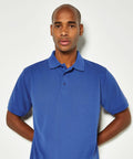 A person wearing a blue short-sleeved Kustom Kit KK403 polo shirt stands in front of a plain white background with hands behind their back.