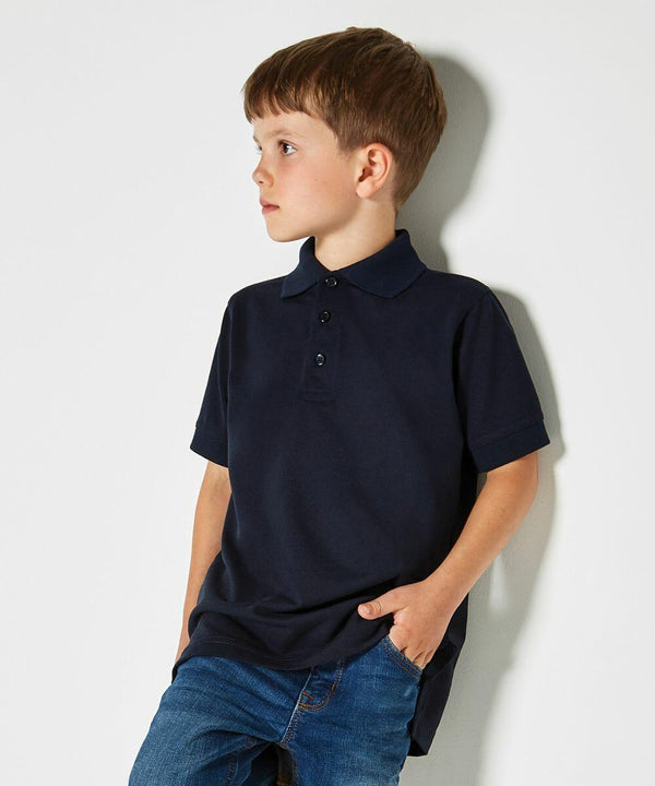 A young boy wearing a navy blue Kustom Kit KK406 polo shirt and blue jeans stands with one hand in his pocket, looking to his left against a plain white background.