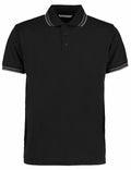 The KK409 by Kustom Kit is a black polo shirt featuring a collar and three buttons, with subtle white stripes adorning the collar and sleeve edges.