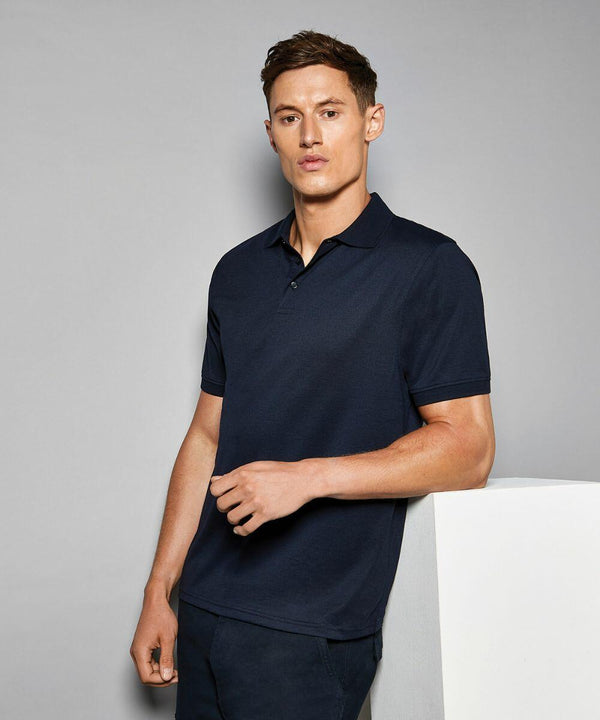 A man in a dark navy Kustom Kit KK412 polo shirt stands against a neutral background, his left hand resting on a white surface.