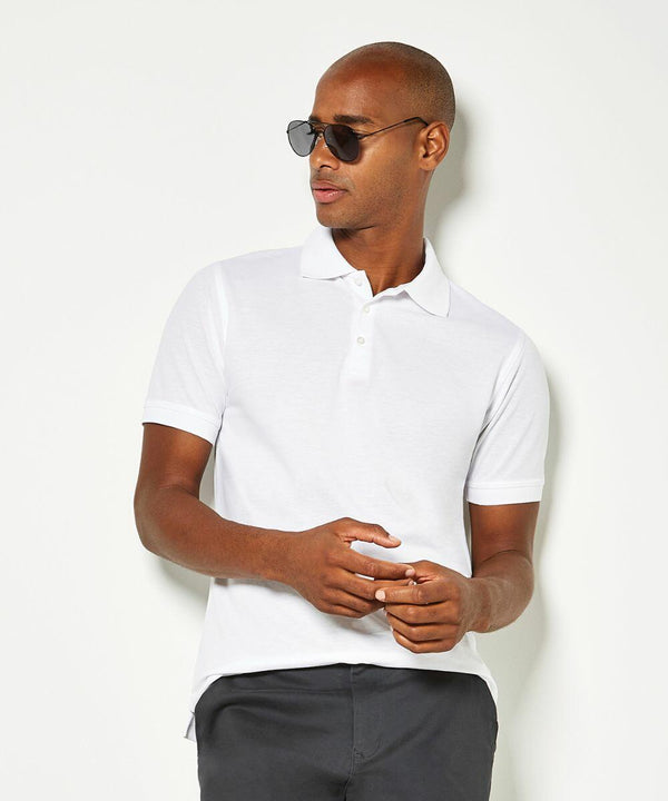 A man wearing a white Kustom Kit KK413 polo shirt, dark sunglasses, and dark pants stands against a plain background.