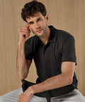 A man with short brown hair and light skin wears a Kustom Kit Regular fit recycled Superwash 60°C polo in black, paired with white trousers, sitting on a black chair against a wooden background, resting his elbow and hand near his face.
