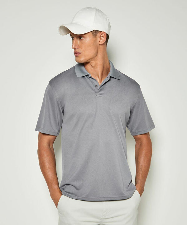 A man wearing a gray Kustom Kit KK444 polo shirt, white cap, and white pants stands against a plain background, looking to his left with hands in his pockets.