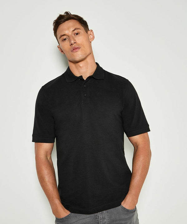 A man stands against a plain white background, wearing a black KK460 short-sleeved polo shirt by Kustom Kit and gray pants.