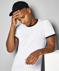 A person wearing a black cap and a white Kustom Kit KK508 t-shirt with black trim rests their head in one hand while leaning on a white surface.