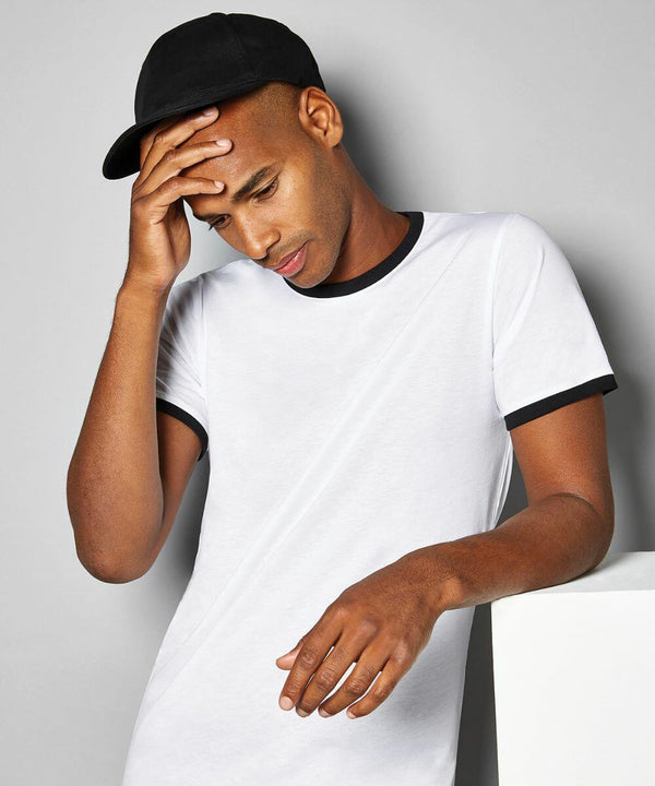 A person wearing a black cap and a white Kustom Kit KK508 t-shirt with black trim rests their head in one hand while leaning on a white surface.