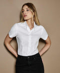 A woman wearing a white Kustom Kit KK701 short-sleeved button-up shirt and black pants stands against a beige background.