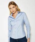 A person with long blonde hair is wearing a light blue KK702 button-up shirt from Kustom Kit and blue jeans, standing against a plain white background.