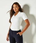 A woman wearing a white short-sleeve KK732 polo shirt by Kustom Kit and dark pants stands against a light background, with one hand in her pocket and hair gently flowing.