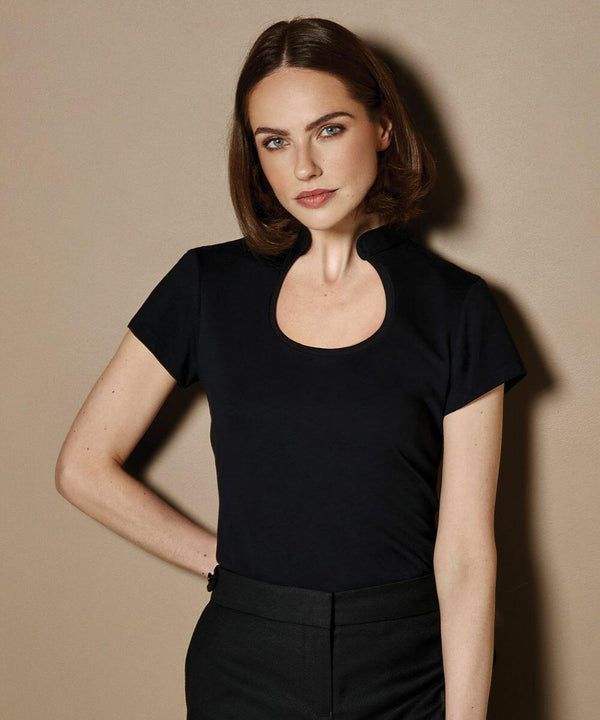 A woman with shoulder-length brown hair, wearing a black short-sleeve KK755 top from Kustom Kit and black pants, stands against a beige background.