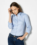 A woman with shoulder-length brown hair, wearing a light blue Kustom Kit KK782 button-up shirt and dark blue jeans, poses against a plain white background.