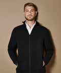 A man with short hair and a beard wears a black zip-up KK902 jacket by Kustom Kit over a white shirt, standing in front of a neutral background with his hands in his pockets.