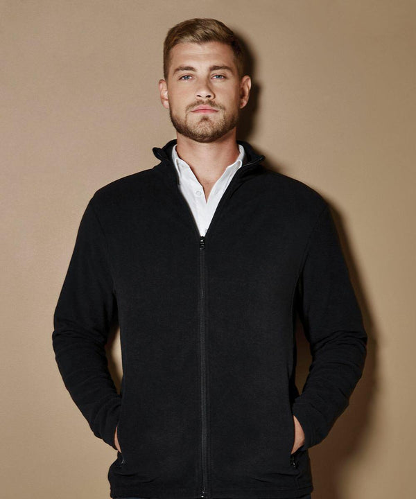 A man with short hair and a beard wears a black zip-up KK902 jacket by Kustom Kit over a white shirt, standing in front of a neutral background with his hands in his pockets.