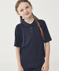 A young girl with light skin and brown plaited hair stands against a plain background, wearing the Finden & Hales Kids team polo shirt in navy blue. She looks at the camera with a slight smile.