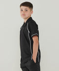A young boy stands sideways, looking at the camera. He wears a Finden & Hales LV372 black polo shirt with white piping and dark pants against a plain background.