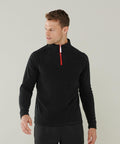 A man stands against a plain background, wearing a Finden & Hales LV570 long-sleeved black pullover with a red zipper. He is looking sideways and has his right hand slightly raised.