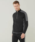 A man stands facing the camera wearing a black and gray Finden & Hales LV571 long-sleeve athletic shirt with a zipper at the collar. He has short brown hair and is smiling slightly.