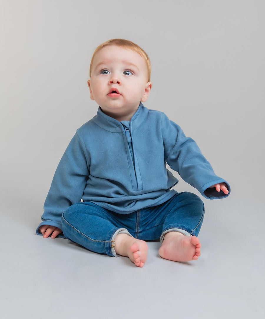A baby with fair hair and blue eyes, barefoot on a light grey floor, wears a blue Larkwood half-zip lightweight fleece pullover and blue jeans, looking slightly upward with a neutral expression.