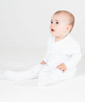 An infant dressed in an all-white LW50T onesie from Larkwood is seated on a white floor, looking upwards with a curious expression.