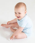 A baby in a light blue Larkwood LW55T sits on the floor, smiling and reaching out with its right hand while its legs are bent.