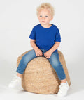 A young child with curly blonde hair wearing a blue t-shirt, jeans, and white sneakers sits on a woven pouf against a plain white background, modeling the LW620 from Larkwood.
