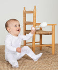 A baby, dressed in a white onesie, sits on a woven mat next to a small wooden chair, holding a white Larkwood LW650.