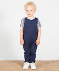 A young child with curly blonde hair stands on a woven mat, wearing a striped shirt under navy blue LW651 overalls by Larkwood, and white sneakers.