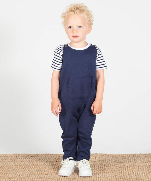 A young child with curly blonde hair stands on a woven mat, wearing a striped shirt under navy blue LW651 overalls by Larkwood, and white sneakers.